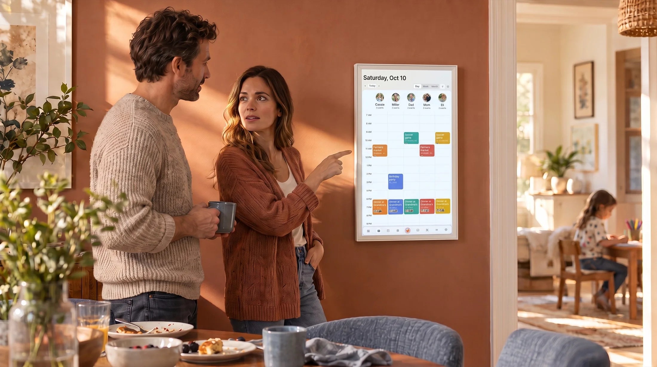 A couple points at the family calendar on the Kora display while their daughter draws at the table